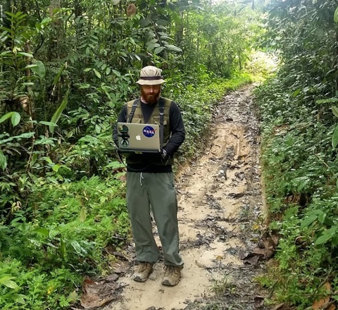 Dr. Bullock standing on a muddy road surrounded by dark green vegetation. He wears a rainhat and a rain jacket, and is holding a laptop computer.
