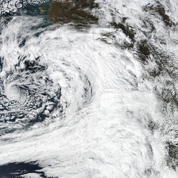 This is a color, visible light image of the atmospheric river over the Gulf of Alaska. On the left side of the image is the blue ocean with the massive white tail and swirl of the atmospheric river spinning counterclockwise over it. To the right is the brown and green of the North American West Coast with clouds over it.