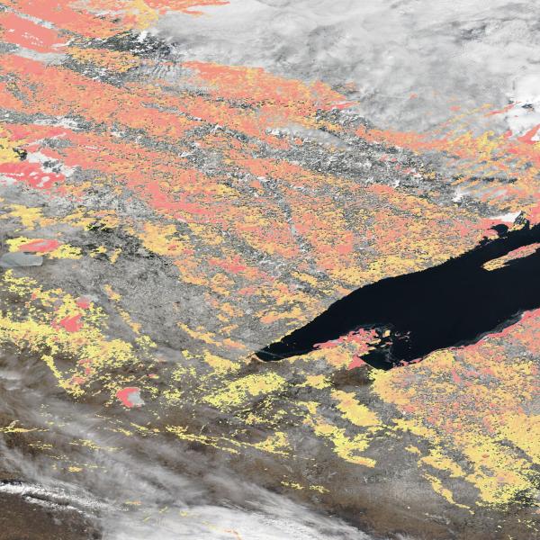 This rectangular image shows snow cover around the Lake Superior region of North America. The base of the image is a color landscape image of the ground with clouds. Overlaid across the image are patches of yellow, orange, and red pixels to mark areas where snow is reflecting light.