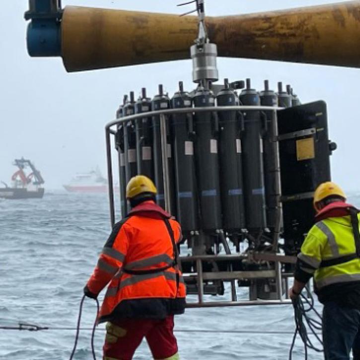 This thumbnail image shows two scientists on a ship preparing to lower a scientific instrument called a rosette into the ocean. The two crewmembers are holding ropes and steadying the instrument. They are wearing orange and green ocean gear and hard hats. The rosette is about six feet tall and consists of many long, gray cylinders oriented in a vertical circle and ringed by a strong, metal frame. 