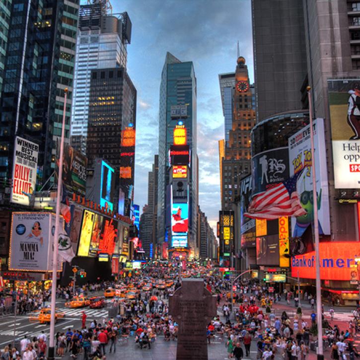 This image features a view of New York City's Times Square looking south in early evening. The right, left, and middle background of the image shows office buildings, skyscapers, and stores with bright lights and advertising signs surrounding the square. The center of the image shows cars and people traversing the streets and sidewalks below.