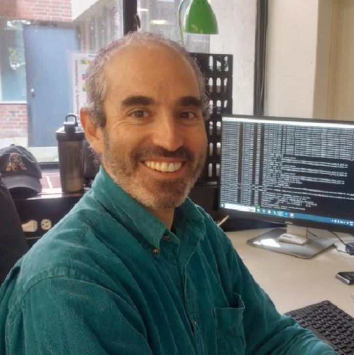 Dr. Gonzalo González Abad, an Atmospheric Physicist at the Center for Astrophysics, sits at a desk in his office. He is wearing a green collared shirt and behind him is a computer monitor showing code used to manipulate data.