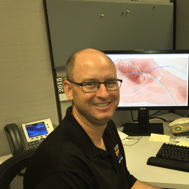Headshot of conway sitting at a desk wearing a dark polo; computer in the background on his desk