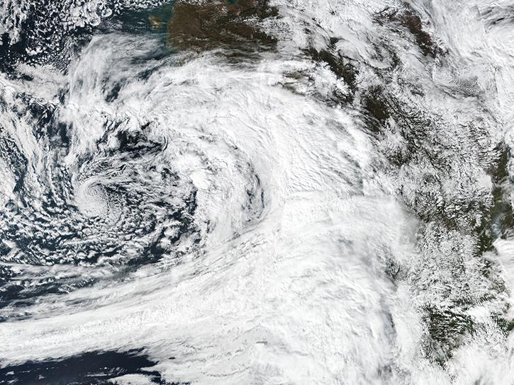 This is a color, visible light image of the atmospheric river over the Gulf of Alaska. On the left side of the image is the blue ocean with the massive white tail and swirl of the atmospheric river spinning counterclockwise over it. To the right is the brown and green of the North American West Coast with clouds over it.