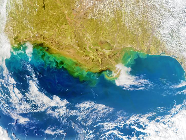 A satellite image of Earth shows blue water, tan land, green shading along the coastline, and wispy white clouds.