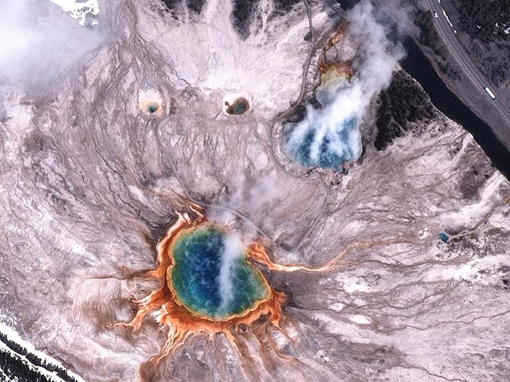 BlackSky Gen-3 satellite image of Grand Prismatic Spring in Yellowstone National Park, Wyoming, at 11:49 a.m. local time on April 21, 2025. A large circular spring (in blue) is surrounded by concentric rings of green, yellow, orange, and red. The landscape consists of pale mineral deposits and patches of snow covered forested areas with dark green trees, and a winding black river. 