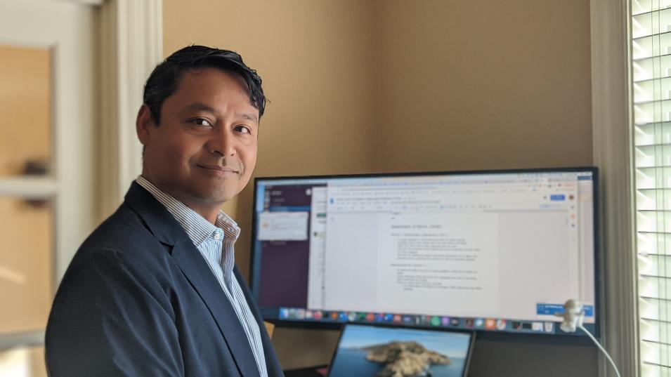 The is a rectangular, landscaped-oriented portrait of Dr. Rahul Ramachandran in a office. Ramachandran is standing on the left side of the image in front of a desk and computer monitor with a window on the right. Ramachandran is wearing a dark sport coat with a light-colored collared shirt.