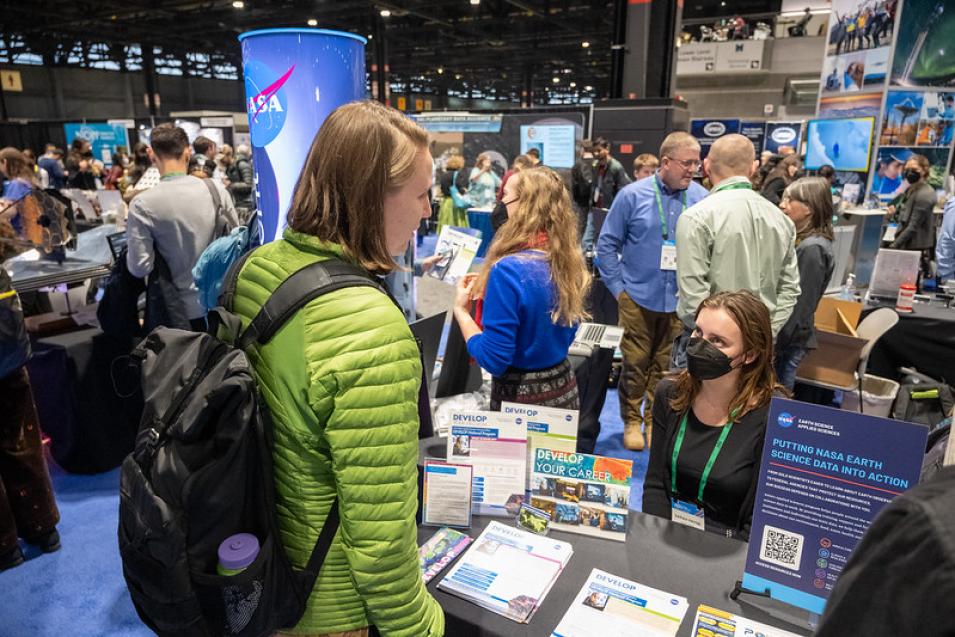 AGU fall meeting attendee in green jacket standing in front of a booth staffed by a sitting NASA staff member