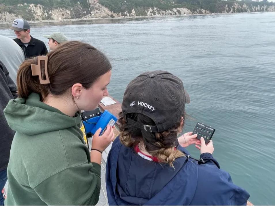 Jillian Ethridge stands in front of the ocean and holds a portable device that tracks ocean color