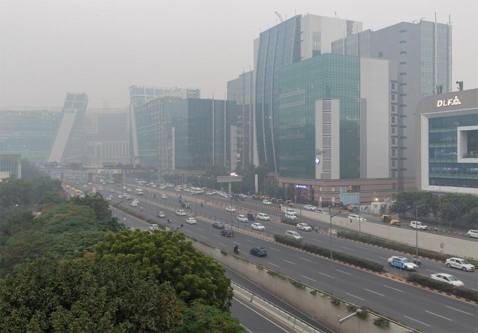 This image shows a busy, smoggy urban scene in Delhi, India. In the background are a line of densely packed together office buildings. Running in front of the buildings is a six-lane boulevard sandwiched in between sideroads with many cars. The air is smoky and the sky is completely grayed out with pollution. 