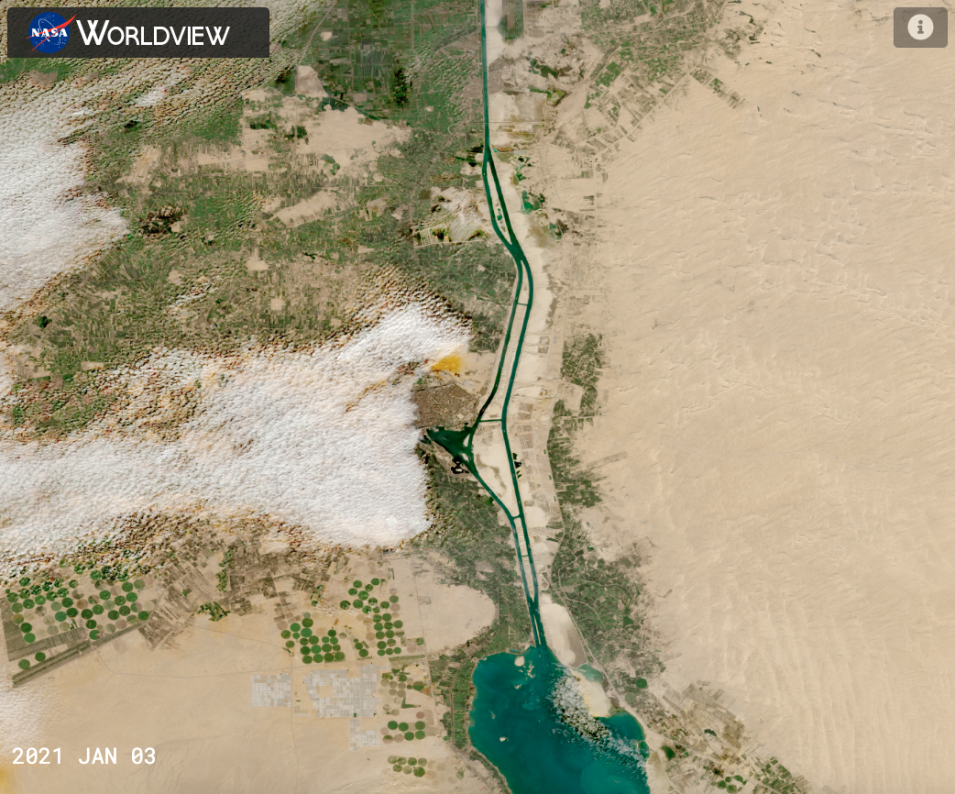 Worldview satellite image of the Suez Canal in Egypt, with the canal shown in bright blue surrounded by tan sand and green brush. A few white clouds appear above land.