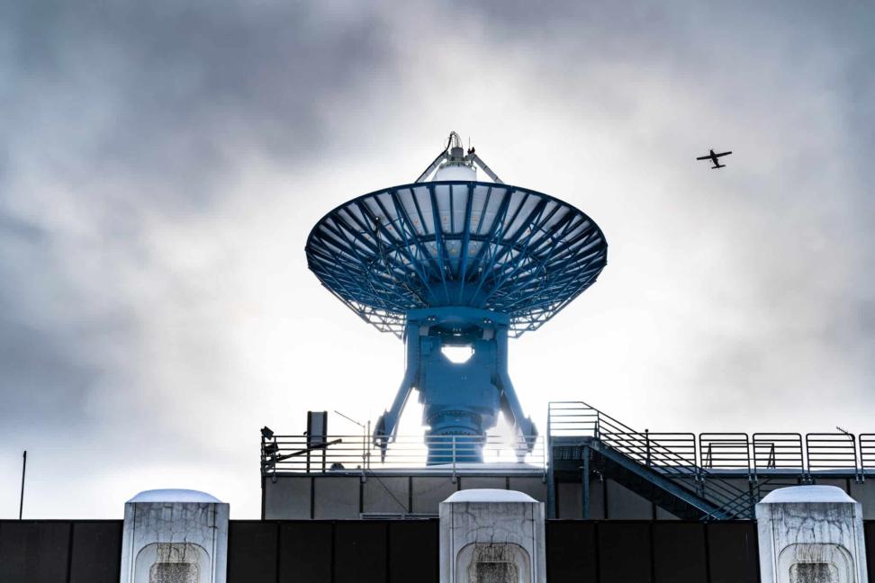 An antenna aimed skyward with an airplane in the sky.