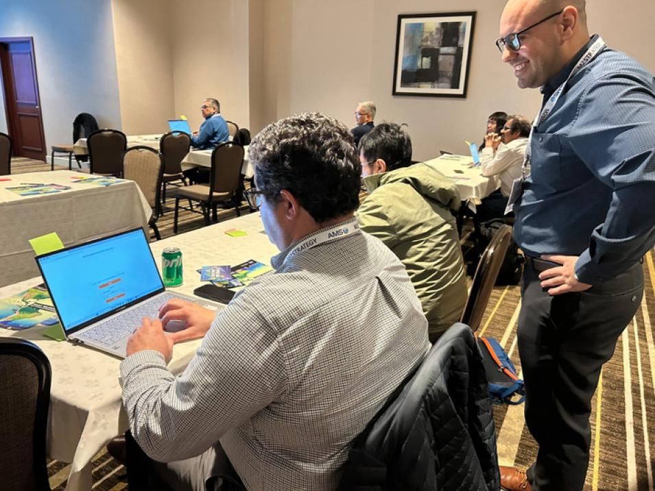 A workshop participant in a white shirt sits in front of an open laptop while a workshop presenter in a blue shirt and glasses looks over his shoulder.