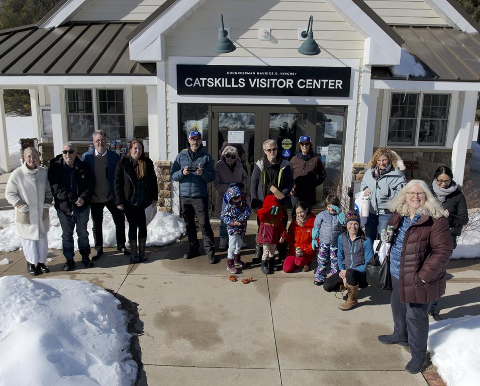 This is a rectangular image of 16 X-Snow participants standing on the sidewalk outside of a white building surrounded by snow. It is a sunny, winter day and the people are wearing warm clothing. On the building is a black sign with white letters reading Catskills Visitor Center.