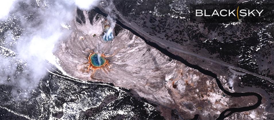 BlackSky Gen-3 satellite image of Grand Prismatic Spring in Yellowstone National Park, Wyoming, at 11:49 a.m. local time on April 21, 2025. A large circular spring (in blue) is surrounded by concentric rings of green, yellow, orange, and red. The landscape consists of pale mineral deposits and patches of snow covered forested areas with dark green trees, and a winding black river. A paved road running parallel to the river and a semi-truck and passenger vehicle can be seen with the BlackSky logo. 