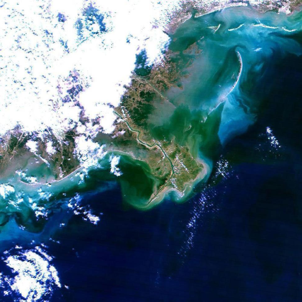 Visible in this square image moving diagonally from the upper left corner to the lower right is a bank of porous clouds, the murky brown and green Louisiana coastline, and deep blue gulf waters.