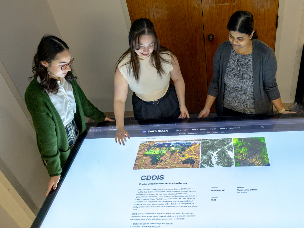 Three people stand around a touch-screen table, which displays information about NASA's CDDIS data archive.