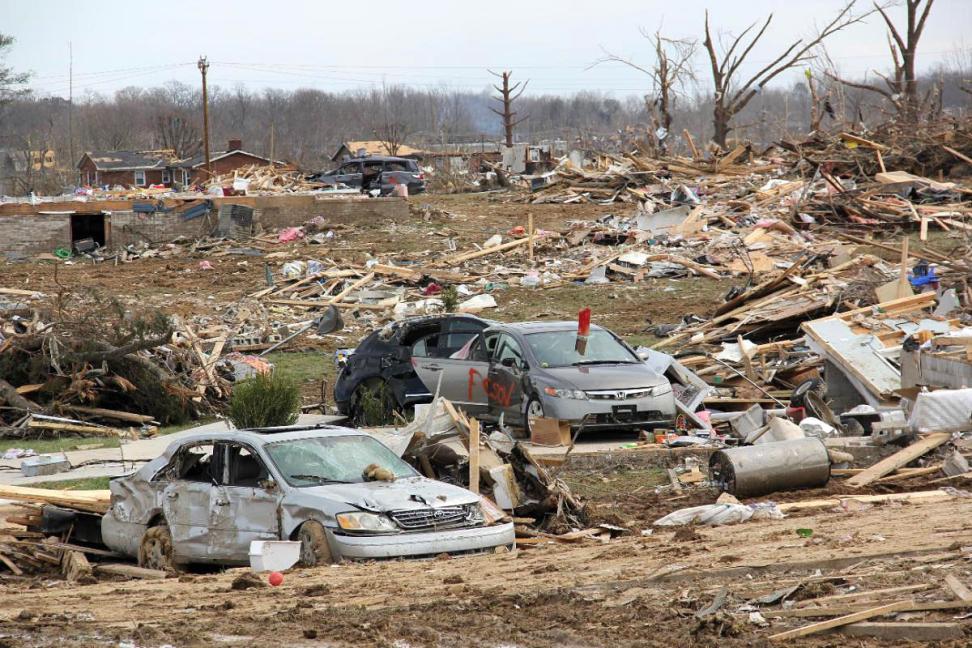 an image of a neighborhood destroyed by a tornado. In the foreground are three smashed cars, and in the background are stripped trees and piles of debris from leveled houses.