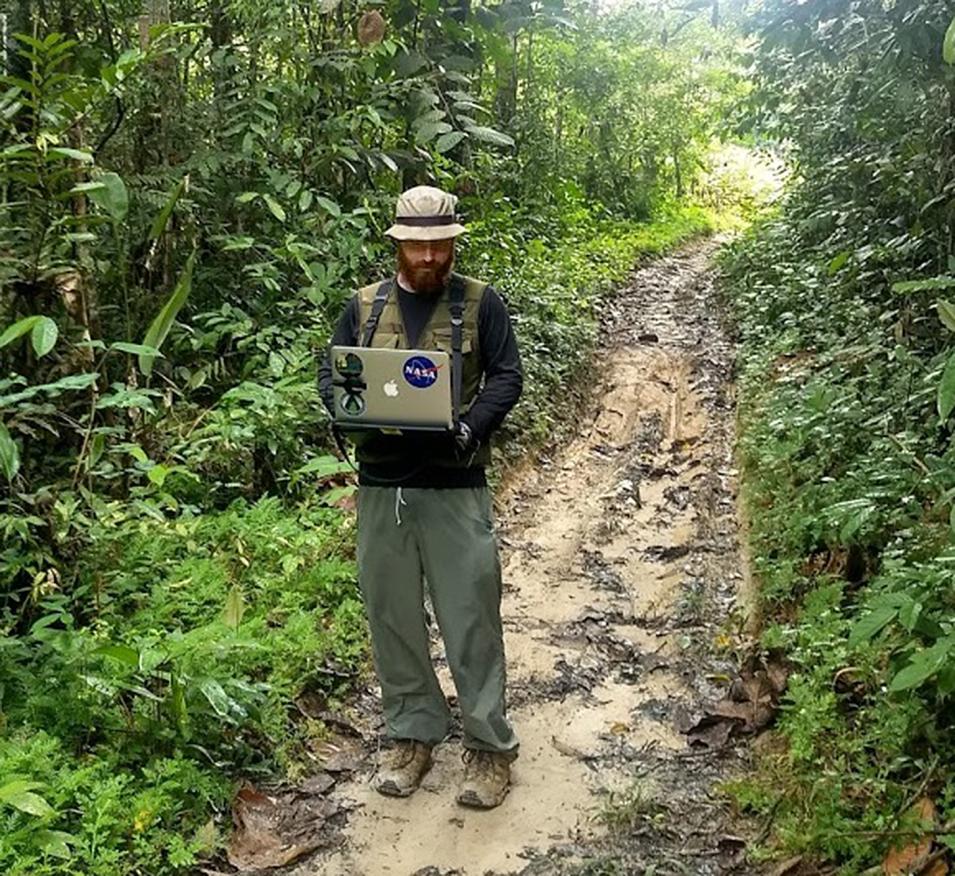 Dr. Bullock standing on a muddy road surrounded by dark green vegetation. He wears a rainhat and a rain jacket, and is holding a laptop computer.