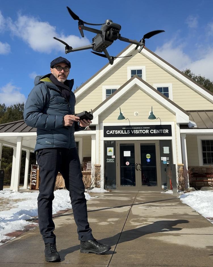 In this rectangular image, a man in winter clothing stands on the sidewalk outside a white building surrounded by snow. The man holds a remote control and is flying a small four-rotor drone a few feet off the ground in front of him. In the background is a blue sky with a few puffy clouds.