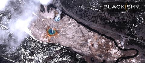 BlackSky Gen-3 satellite image of Grand Prismatic Spring in Yellowstone National Park, Wyoming, at 11:49 a.m. local time on April 21, 2025. A large circular spring (in blue) is surrounded by concentric rings of green, yellow, orange, and red. The landscape consists of pale mineral deposits and patches of snow covered forested areas with dark green trees, and a winding black river. A paved road running parallel to the river and a semi-truck and passenger vehicle can be seen with the BlackSky logo. 