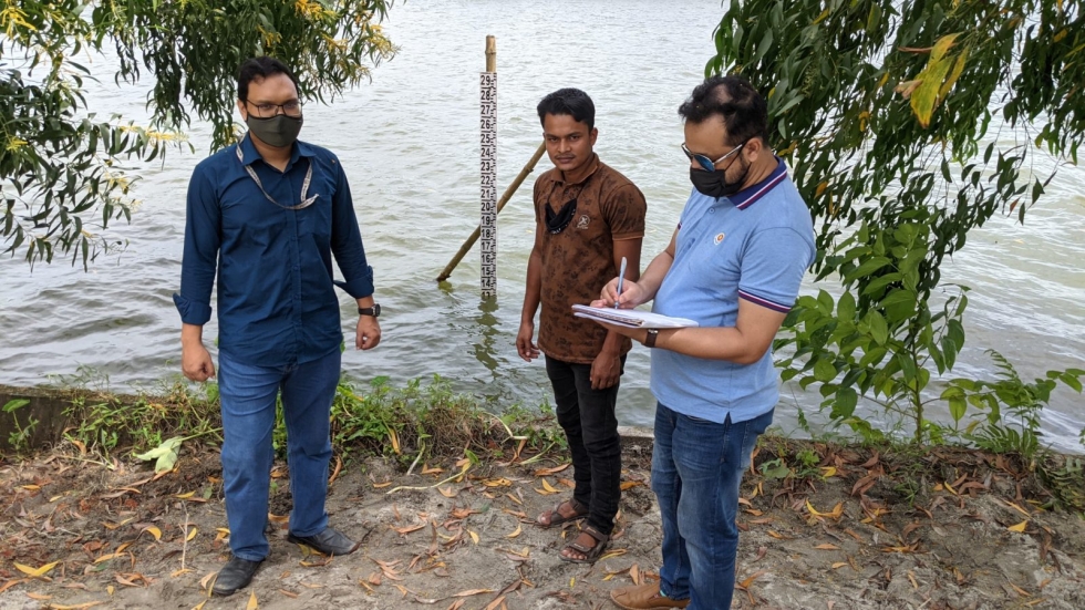 Three citizen scientists standing on the bank of a lake. A lake height gauge is in the background and one man is writing data in a book.