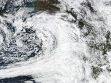 This is a color, visible light image of the atmospheric river over the Gulf of Alaska. On the left side of the image is the blue ocean with the massive white tail and swirl of the atmospheric river spinning counterclockwise over it. To the right is the brown and green of the North American West Coast with clouds over it.