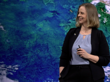 This is an image of NASA's Earth Data Officer, Katie Baynes, standing in front of a blue and green image of Earth. She has a black sweater on with a gray shirt underneath and black pants.