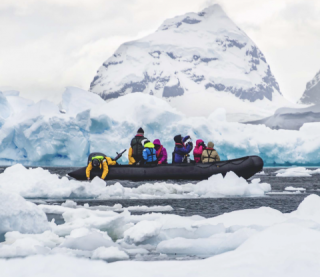 Seven volunteers dressed in colorful drysuits ride in a gray inflatable boat moving through waters containing floating sea ice. In the background is cloudy sky framing a large snow-covered iceberg, rock or land formation.