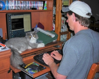 Image of Dr. Thome working at his home computer with his back to the camera. His gray and white cat Aster is sitting in front of the computer facing the camera.